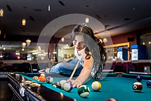 Brunette is sitting on table and playing billiards.
