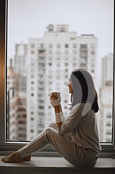 Brunette sitting on a windowsill and drinks a coffee