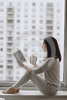 Brunette sitting on a windowsill and drinks a coffee