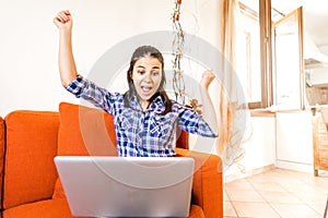 Brunette smart gen z girl rejoices in front of with open raised arms sitting at home sofa. Shocked young woman looking at computer