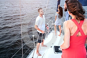 Brunette stands on bow of yacht and looks down. She poses. Two young men looking and talking with each other. Readhead