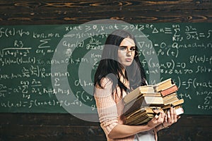 Brunette student walking with two heaps of books in front of green board full of writing. Math undergraduate preparing photo