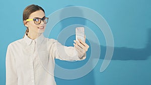 Brunette is Taking Selfie by Smartphone on Blue Background in Studio. Attractive Hipster Girl Wearing Fashionable
