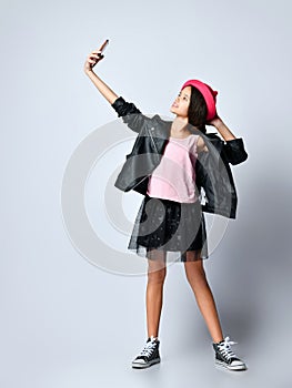Teenage kid in black leather jacket, skirt and pink hat. She taking selfie using smartphone, posing isolated on white. Close up