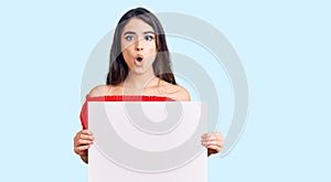 Brunette teenager girl holding blank empty banner scared and amazed with open mouth for surprise, disbelief face