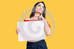 Brunette teenager girl holding blank empty banner serious face thinking about question with hand on chin, thoughtful about