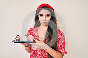 Brunette teenager girl holding cake sweets thinking attitude and sober expression looking self confident