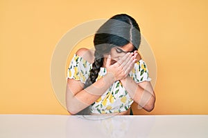 Brunette teenager girl wearing casual clothes sitting on the table with sad expression covering face with hands while crying