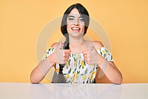 Brunette teenager girl wearing casual clothes sitting on the table success sign doing positive gesture with hand, thumbs up