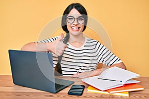 Brunette teenager girl working at the office with laptop smiling happy and positive, thumb up doing excellent and approval sign