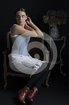 A Victorian woman wearing a corset and combinations and sitting on a chair in front of a dark backdrop