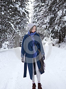 Brunette walks through the winter forest in a gray hat, blue sweater and blue coat.