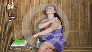 A brunette woman in a bath in a towel smears the body with moisturizing milk.
