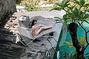 Brunette woman in beige bikini relaxing lying on lounge chair at the poolside near luxury pool in tropic on vacation