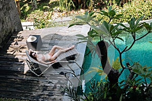 Brunette woman in beige bikini relaxing lying on lounge chair at the poolside near luxury pool in tropic on vacation