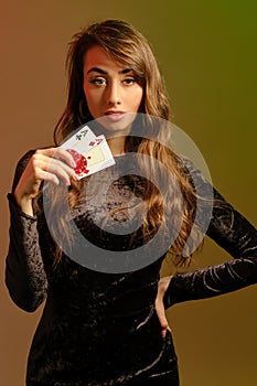 Brunette woman in black dress showing two red chips and aces, posing against colorful studio background. Gambling, poker