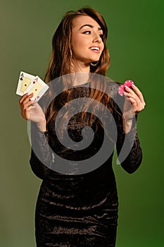Brunette woman in black dress is smiling, showing two red chips and aces, posing against colorful studio background