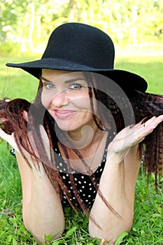 Brunette woman in black hat lies on grass in park on belly leaning on elbows. leaning his head on his hands, looks in