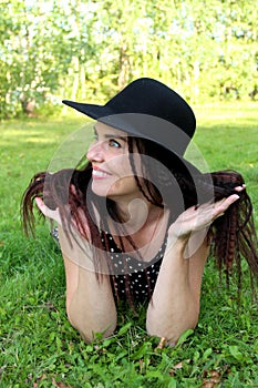 Brunette woman in black hat lies on grass in park on belly leaning on elbows. leaning his head on his hands, looks away