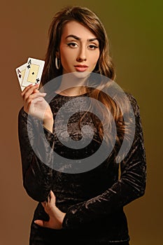 Brunette woman in black velvet dress and jewelry showing two aces, posing against colorful studio background. Gambling