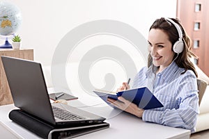 Brunette woman in blue shirt with headphones at laptop makes notes in notebook, home-schooled, online courses, remote work at home photo