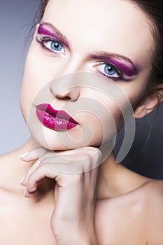 Brunette woman with creative make up violet eye shadows full red lips, blue eyes and curly hair with her hand on her face