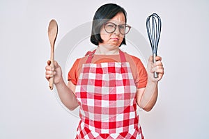 Brunette woman with down syndrome cooking using baker whisk and spoon skeptic and nervous, frowning upset because of problem