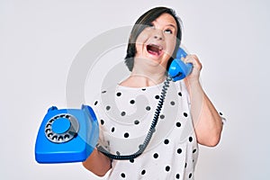 Brunette woman with down syndrome holding vintage telephone angry and mad screaming frustrated and furious, shouting with anger
