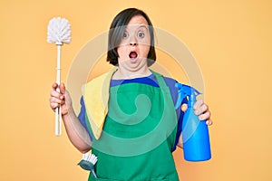 Brunette woman with down syndrome wearing apron holding scourer and toilet brush afraid and shocked with surprise and amazed