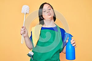 Brunette woman with down syndrome wearing apron holding scourer and toilet brush looking at the camera blowing a kiss being lovely