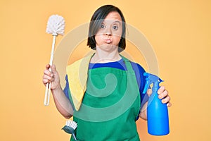 Brunette woman with down syndrome wearing apron holding scourer and toilet brush puffing cheeks with funny face