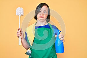 Brunette woman with down syndrome wearing apron holding scourer and toilet brush relaxed with serious expression on face