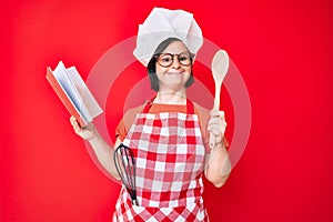 Brunette woman with down syndrome wearing professional baker apron reading cooking recipe book smiling with a happy and cool smile