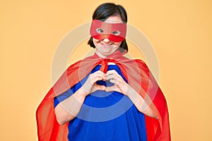 Brunette woman with down syndrome wearing super hero costume smiling in love showing heart symbol and shape with hands