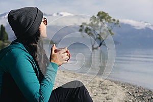 Brunette woman drinking coffee in Wanaka Lake admiring iconic tree on the water. New Zealand, South Island