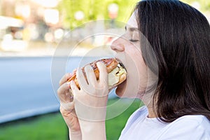 Brunette woman eating burger on the street near the road in the city. Fast food on the way to work or school. Place for