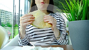 Brunette woman eats pita sandwich with cheese and herbs in a cafe, sitting at a table near the window.