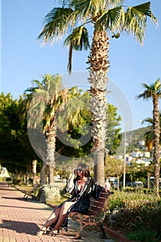 A brunette woman with glasses is sitting on a bench on the street near palm trees