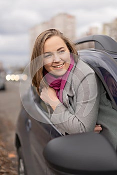 Brunette woman half get out through car window in autumn coat. She smiling cheerfully