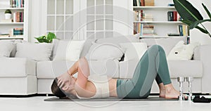 Brunette woman is having abdominal press exercises. She is doing it on the mat next to the sofa at home in living room