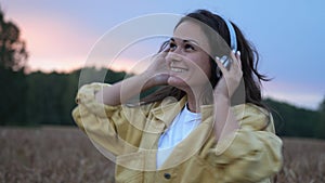 Brunette woman in headphones dancing in wheat field in summer sunset time