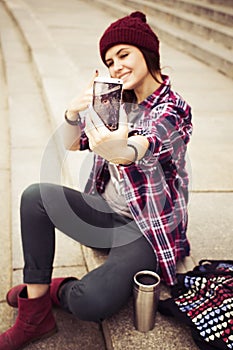 Brunette woman in hipster outfit sitting on steps and taking selfie on phone on the street. Toned image