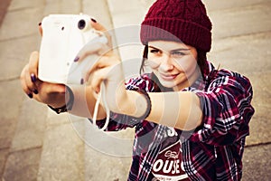 Brunette woman in hipster outfit sitting on steps and taking selfie on retro camera on the street. Toned image