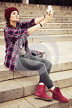 Brunette woman in hipster outfit sitting on steps and taking selfie on retro camera on the street. Toned image