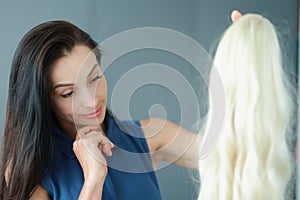 Brunette woman holding blonde wig with long hair and thinking to wear it