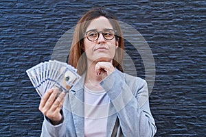 Brunette woman holding dollars serious face thinking about question with hand on chin, thoughtful about confusing idea