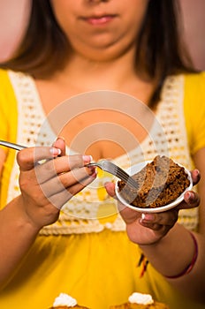 Brunette woman holding small plate of chocolate brownie cake, grabbing bite using fork, showing to camera, pastry