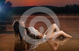 brunette woman in lingerie laying in river water. Young female relaxing on the beach during sunset. Perfect body girl