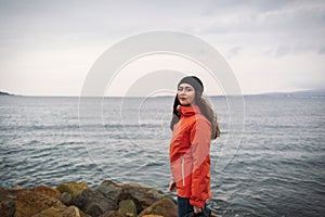 Brunette woman with long hair in a hat and an orange jacket, posing on the beach. In the background the sea and the horizon line.