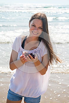 Brunette woman with long hair in shorts and light jacket on beach. Holds cherry in her hands. Smile